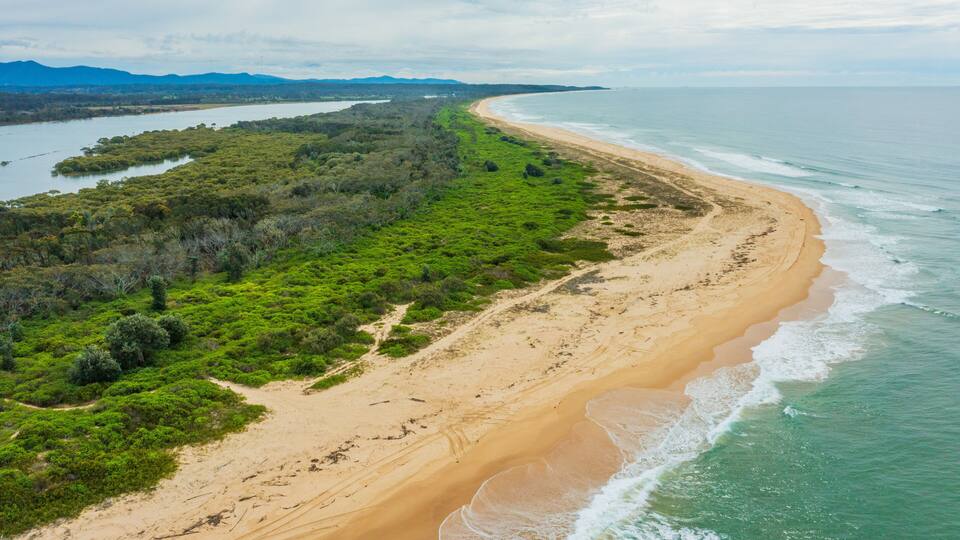 A coastal landscape with a sandy beach, dense green vegetation, and a river or bay.