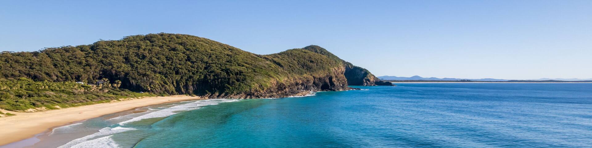 A coastal landscape with clear turquoise waters, a sandy beach, and a forested hill.