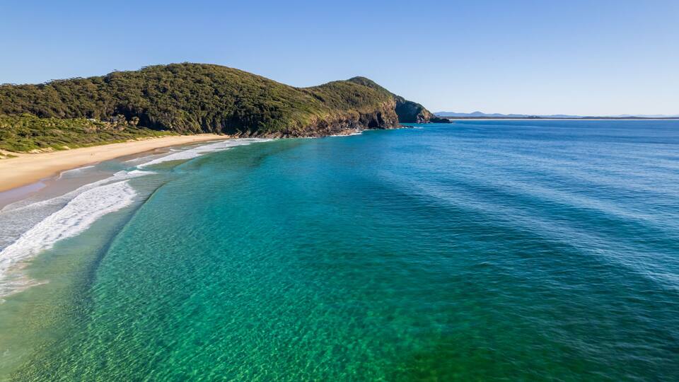 A coastal landscape with clear turquoise waters, a sandy beach, and a forested hill.