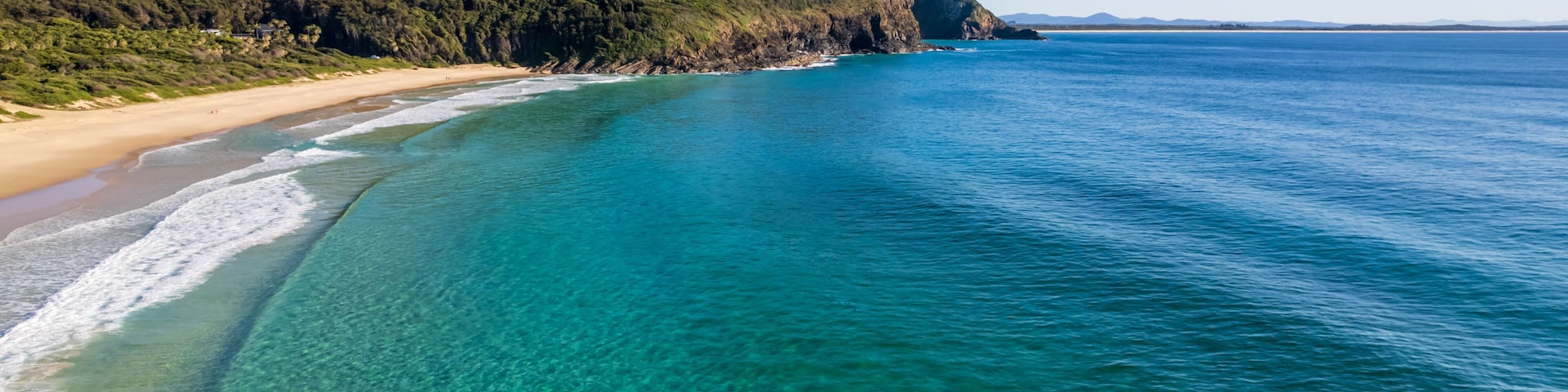 Paisagem costeira com águas turquesa cristalinas, uma praia arenosa e uma colina arborizada.