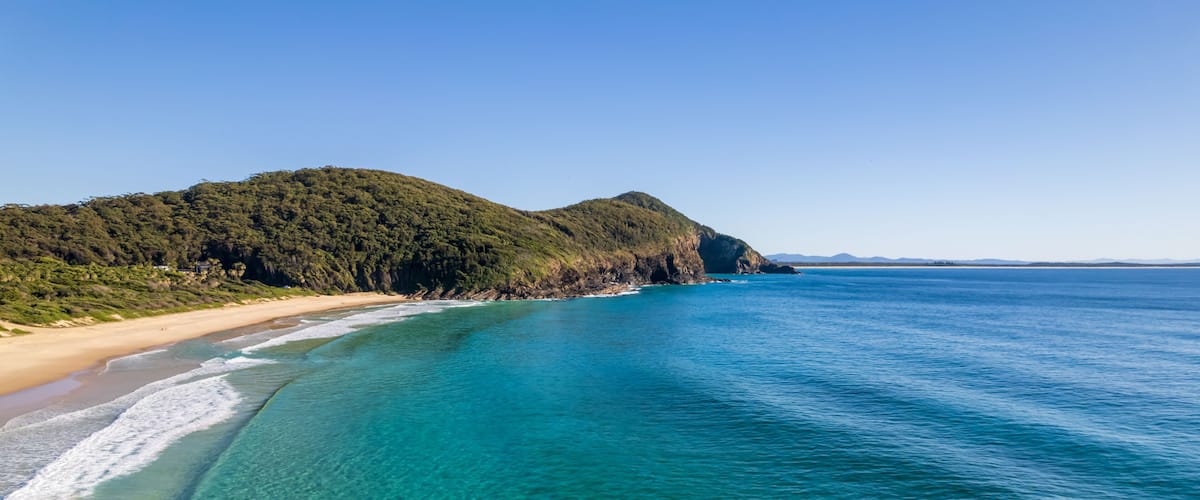 A coastal landscape with clear turquoise waters, a sandy beach, and a forested hill.