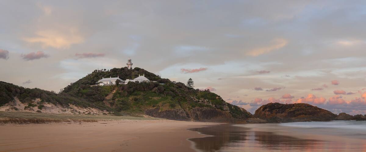 A beach with a white lighthouse on a hill, rocky outcrops, and a sunset sky.