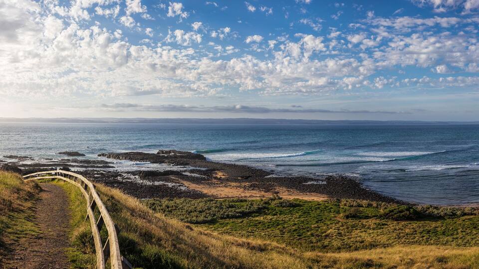 A coastal landscape with a curving road, rocky shoreline, and grassy hills.