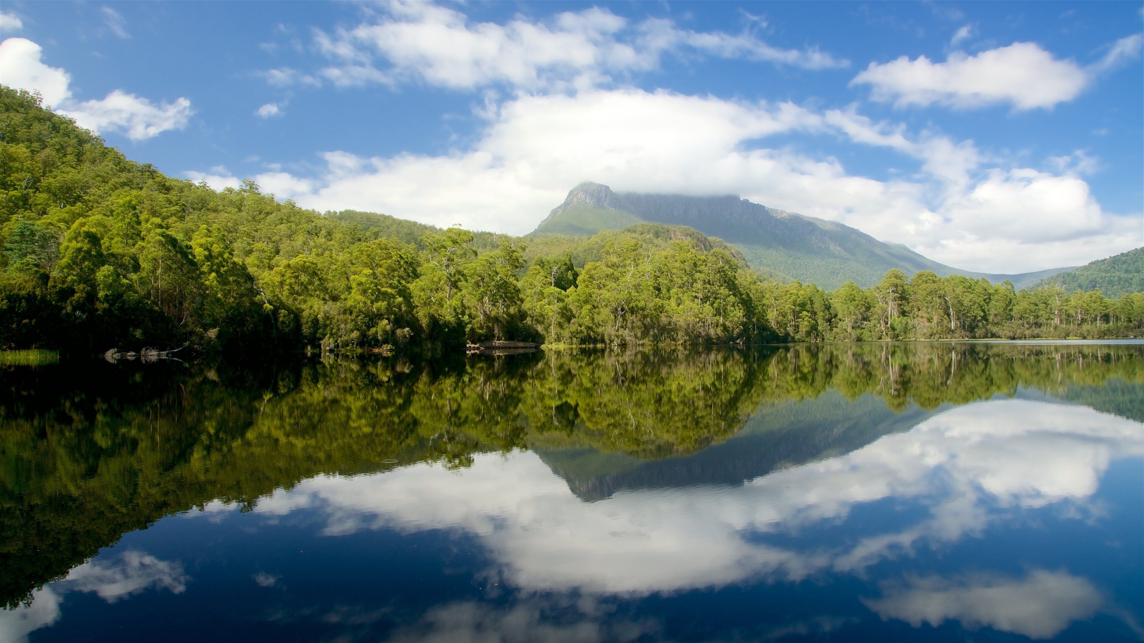 A calm lake reflecting trees and mountains under a partly cloudy sky.