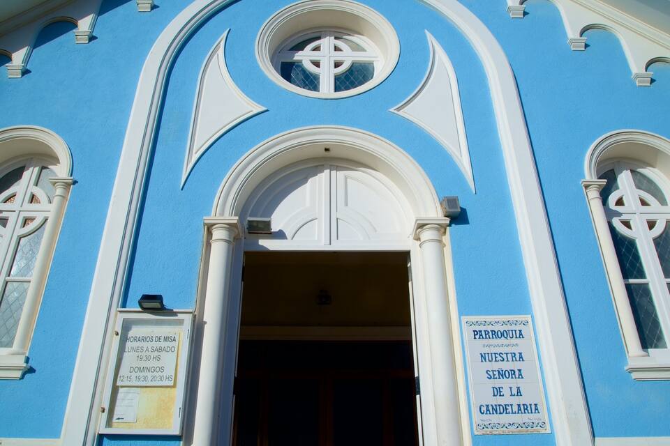 Edifício azul com detalhes brancos e janelas em arco. Há uma placa na porta que diz “Horarios de Misá” com horários listados de segunda a sábado.