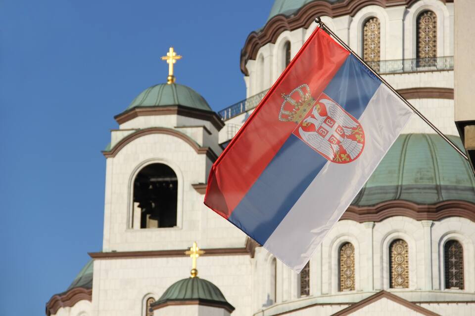 A flag with a red, white, and blue design with a crown and a shield, in front of a church with domed architecture and a cross on top.