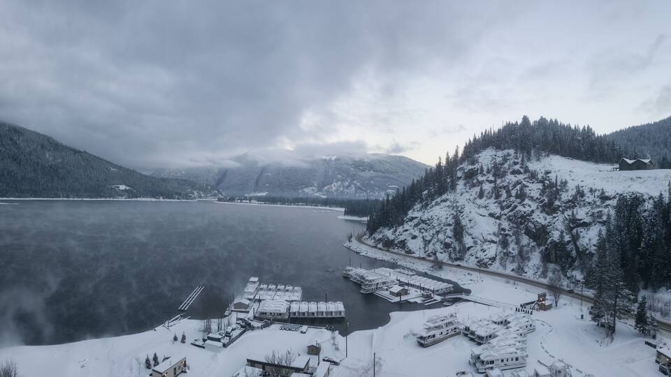 A snow-covered lake with docked boats, a dock, and a cabin on a snowy hillside.