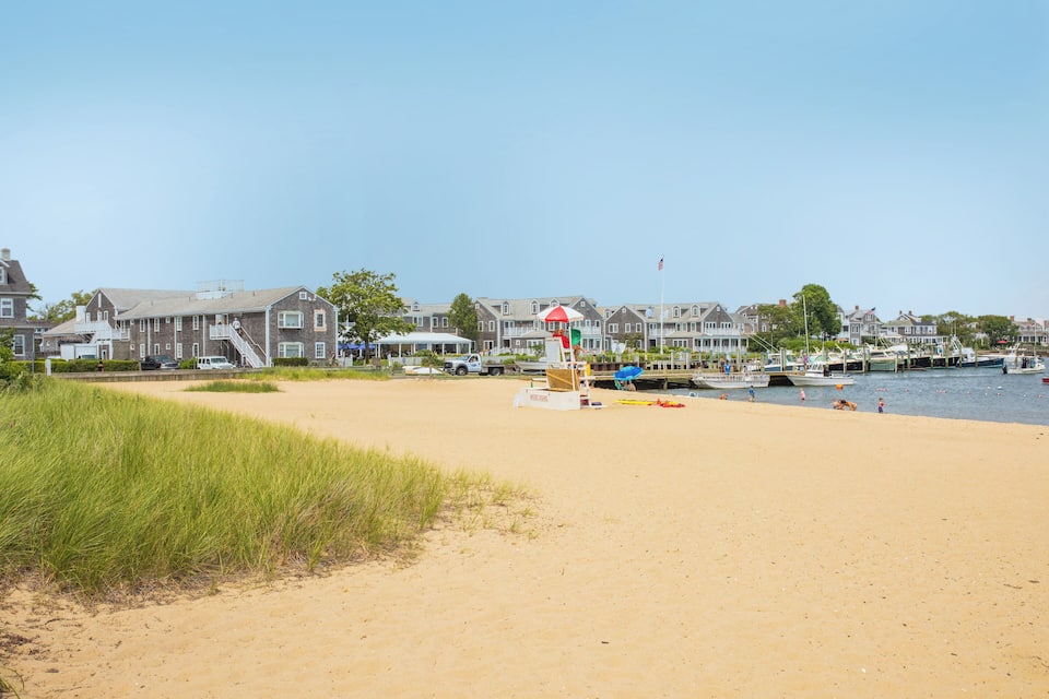 A sandy beach with a lifeguard tower, boats, and houses in the background.