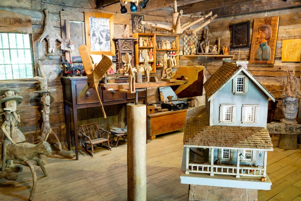 A rustic wooden room with various wooden sculptures, a small model house, and a sign that reads "EXHIBITS".