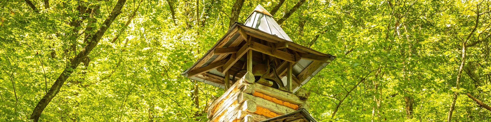 A wooden cabin with a steeply pitched roof and a chimney, surrounded by dense green foliage.