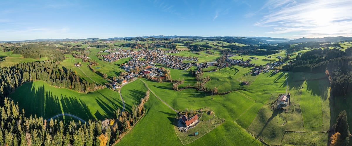 A panoramic view of a rural town nestled in rolling hills, surrounded by lush green fields and dense forests.
