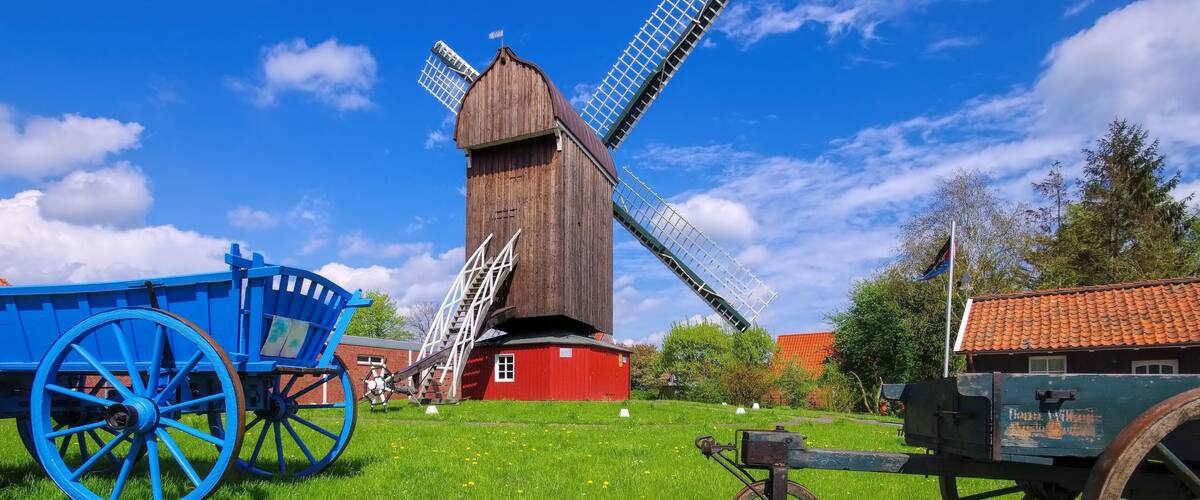 A traditional windmill with a red base and a wooden structure, surrounded by green grass and a blue cart.