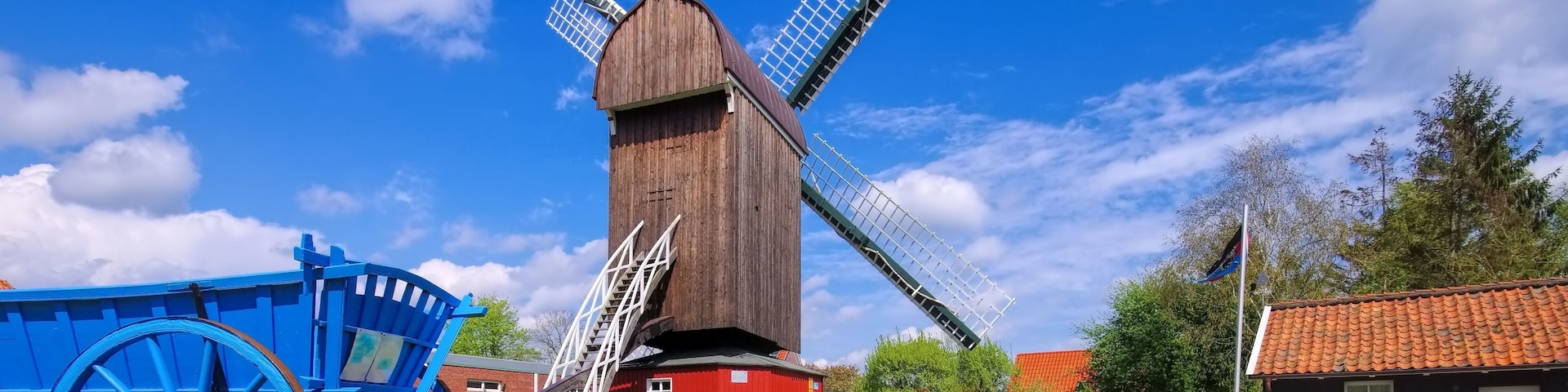 A traditional windmill with a red base and a wooden structure, surrounded by green grass and a blue cart.