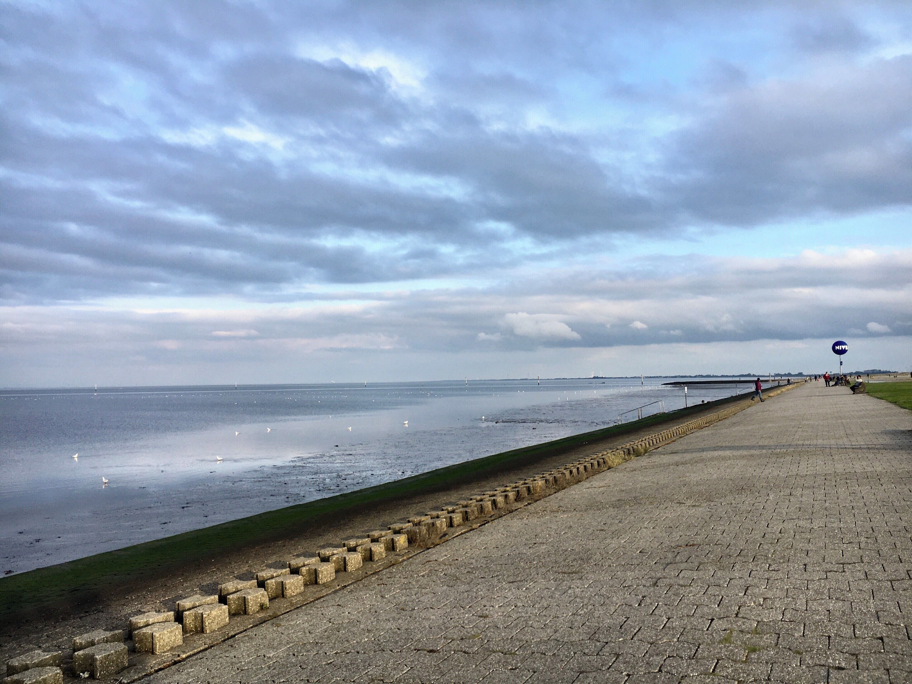 A coastal walkway with a stone barrier, a grassy area, and a body of water with sailboats.