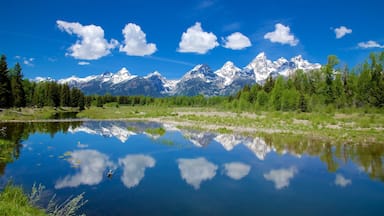 Uma cordilheira com picos nevados, céu azul límpido e um lago tranquilo refletindo a paisagem.