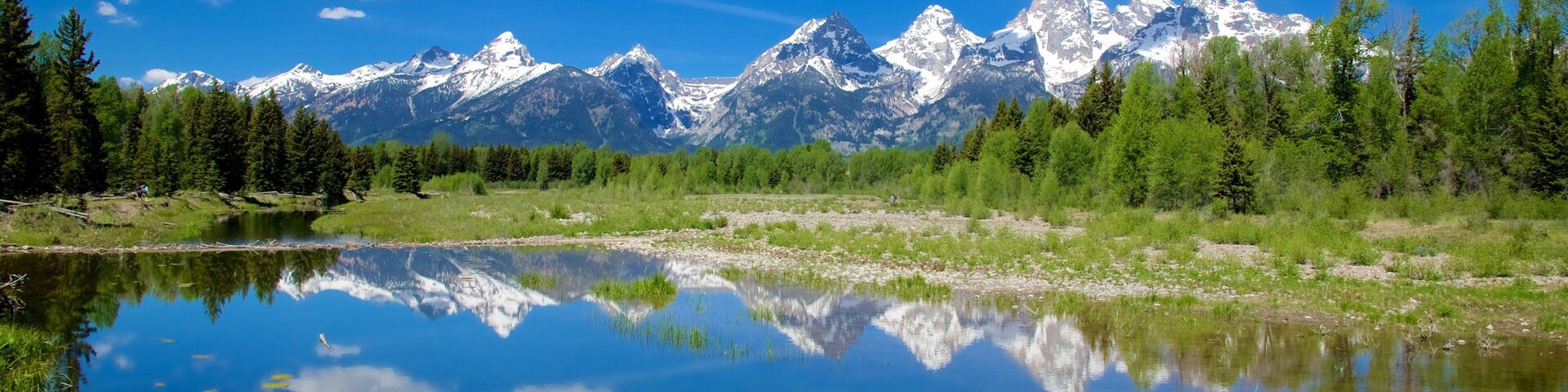 Cordillera con picos nevados, cielo azul despejado y un lago tranquilo que refleja el paisaje.