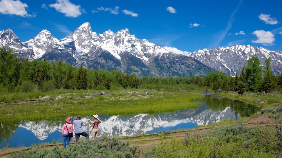 A mountain range with snow-capped peaks, a calm lake reflecting the scenery, and a group of people by the water.