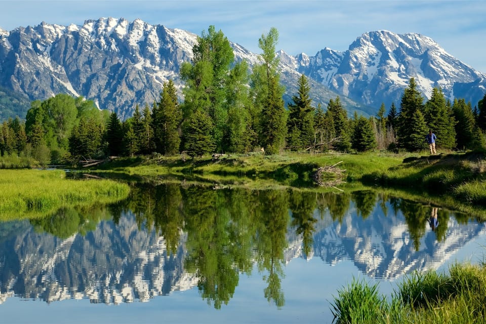 A mountain range with snow-capped peaks, a calm lake reflecting the trees, and a person hiking in the foreground.