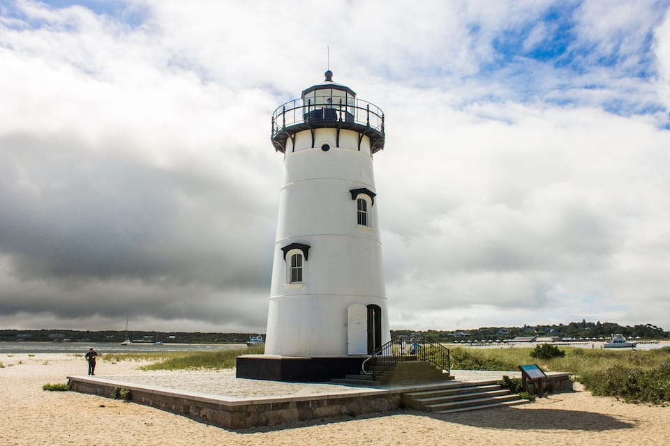 Een witte vuurtoren met een zwarte bovenkant, gelegen op een zandstrand met een grasveld op de voorgrond en een bewolkte lucht op de achtergrond.