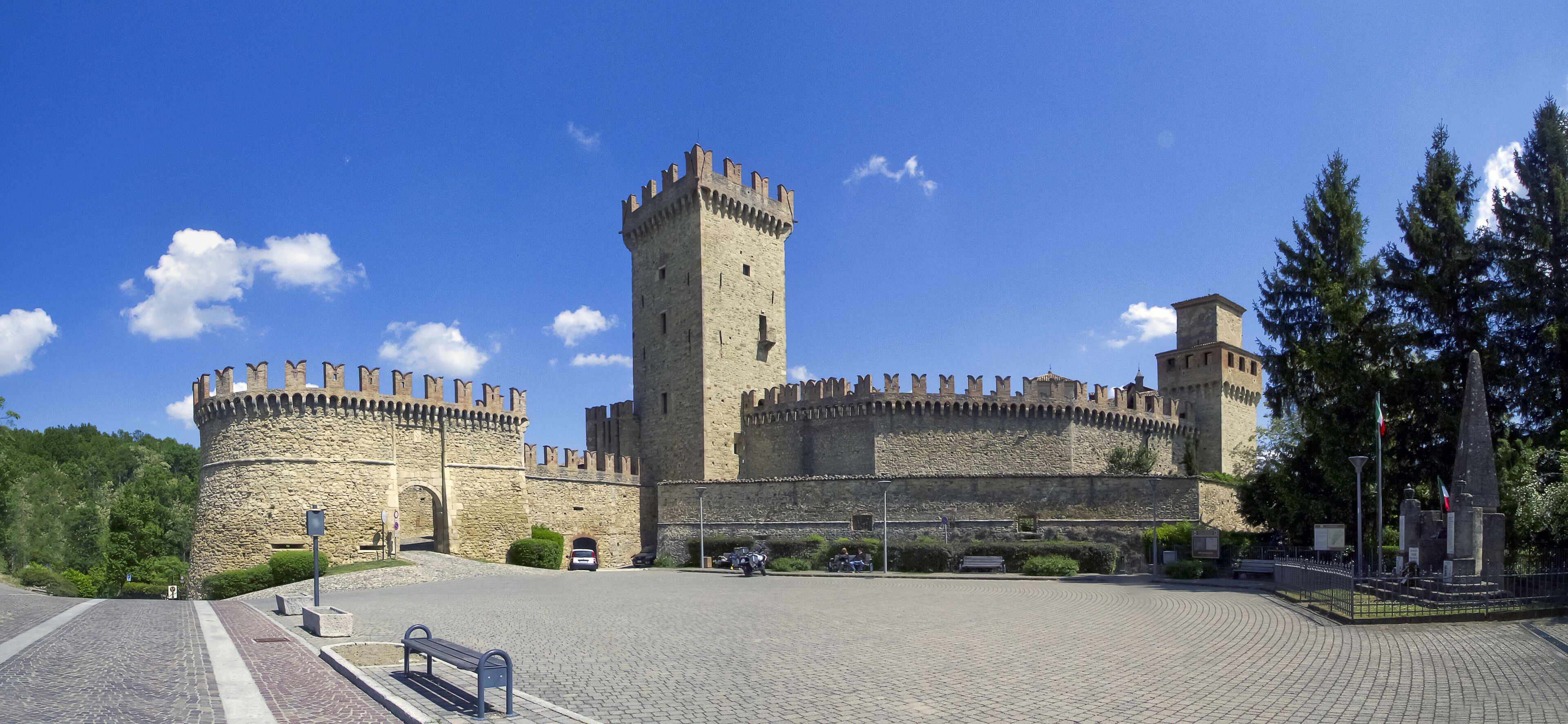 A historic stone castle with a central tower and two round towers, surrounded by a paved courtyard and greenery.