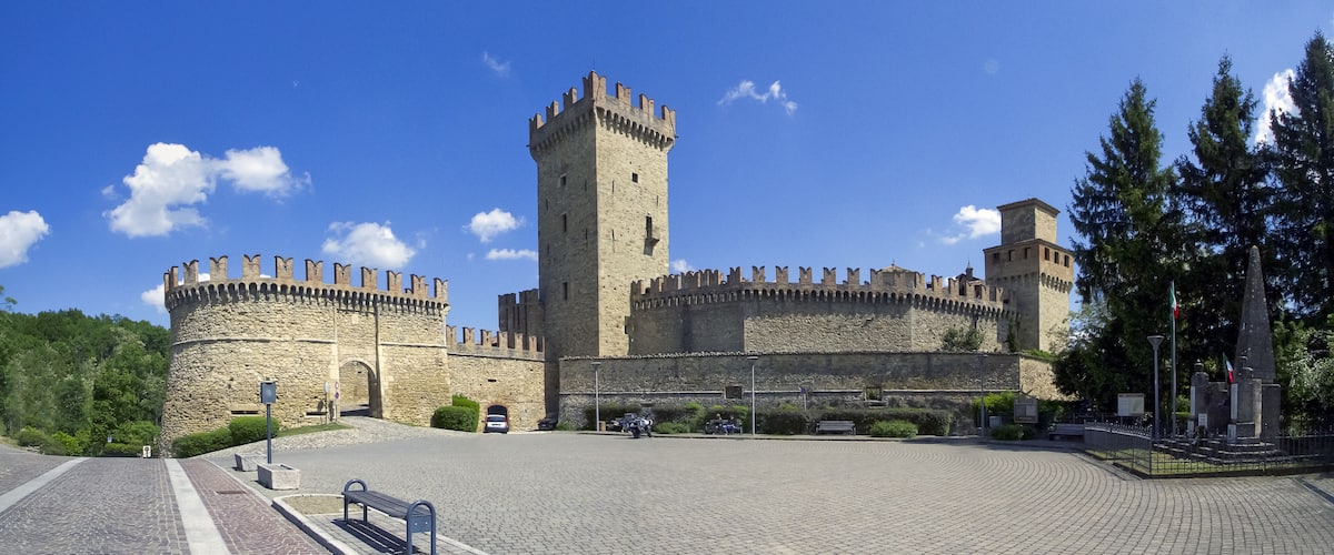 A historic stone castle with a central tower and two round towers, surrounded by a paved courtyard and greenery.