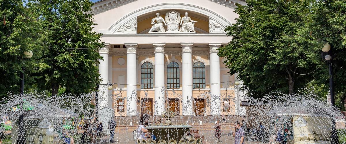 A classical building with columns and statues, surrounded by a fountain and trees.
