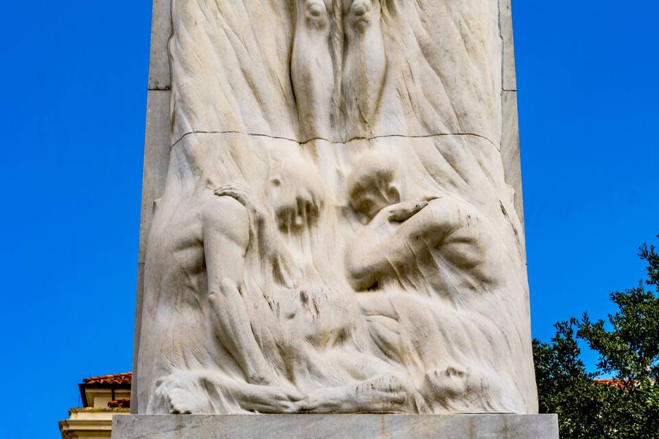 Uma grande escultura de mármore retratando um homem deitado com os braços estendidos, em frente a um céu azul claro.