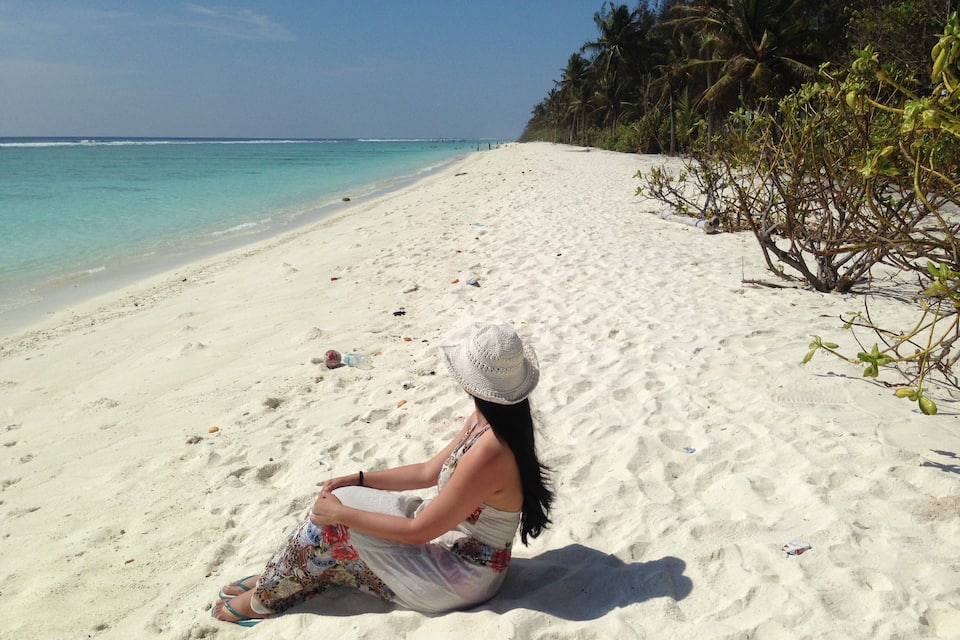 A woman sitting on a sandy beach with turquoise water and palm trees in the background.