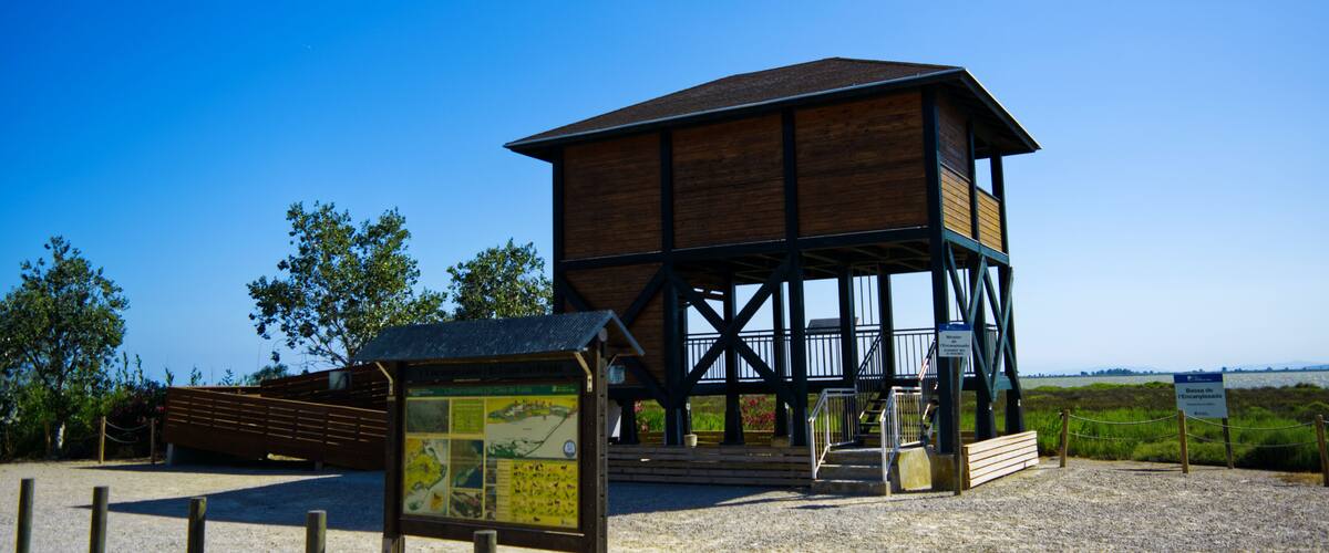 A wooden observation tower with a metal staircase, surrounded by a gravel area and a signboard.