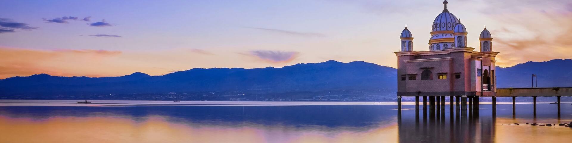 A coastal scene with a historic building on stilts, a calm sea, and mountains in the background.