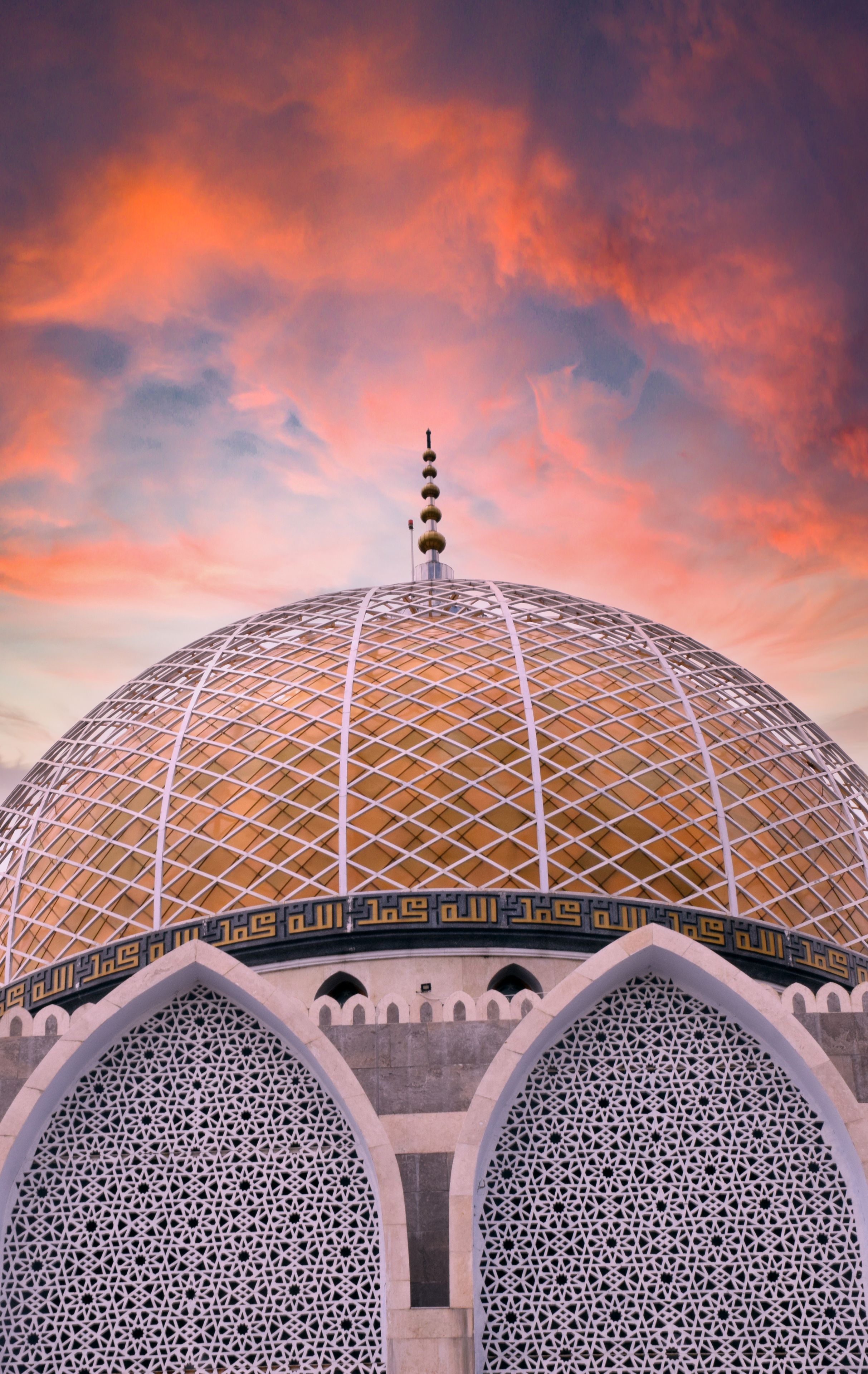 A large, domed structure with intricate geometric patterns and Arabic calligraphy, set against a vibrant sunset sky.