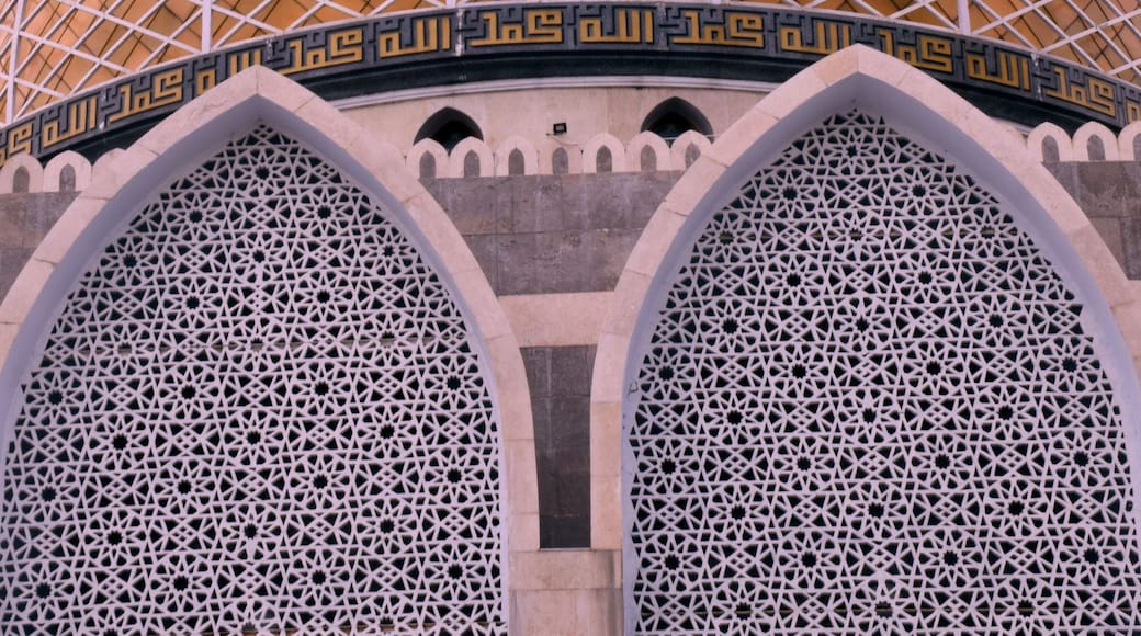 A large, domed structure with intricate geometric patterns and Arabic calligraphy, set against a vibrant sunset sky.