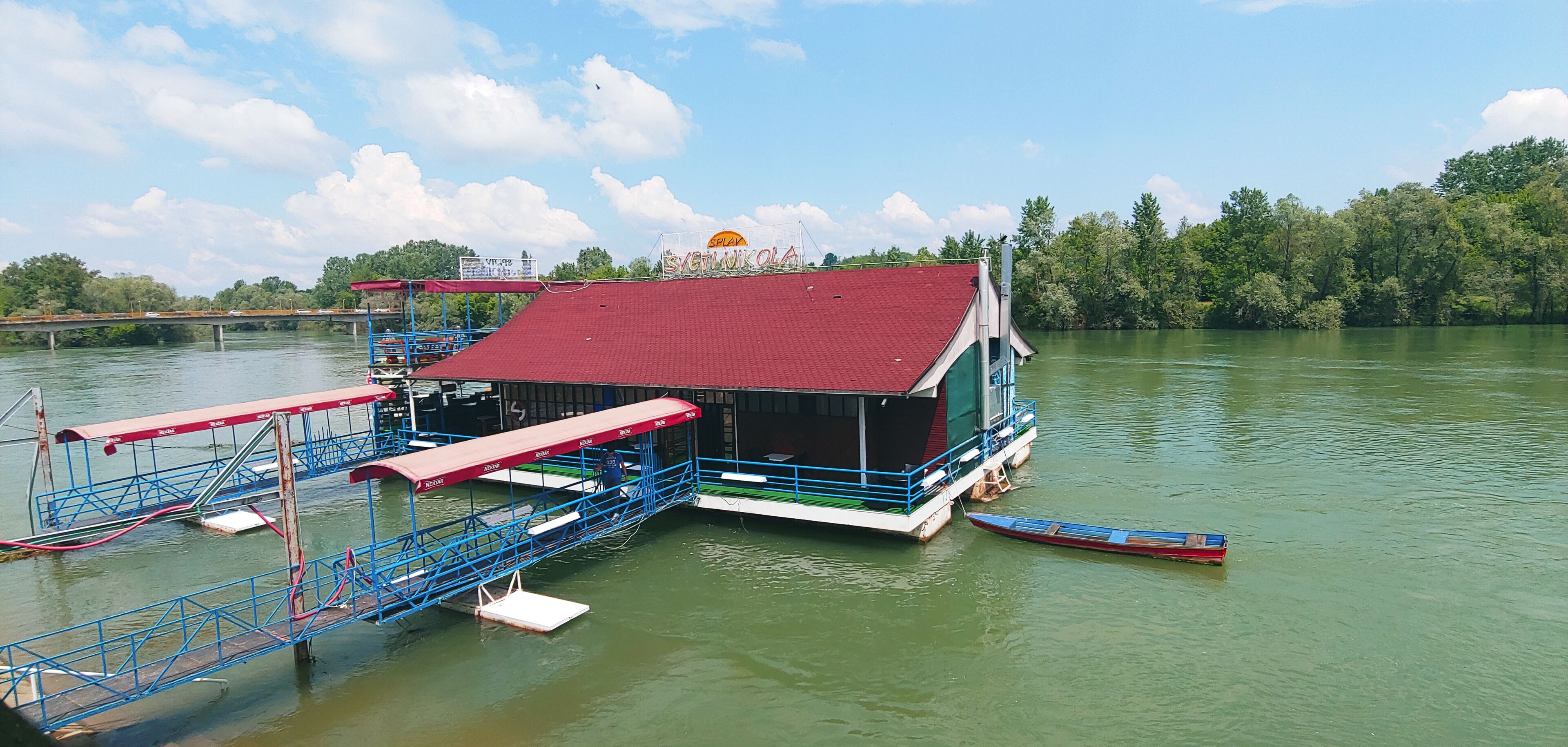 A floating house with a red roof and a sign that reads "Svetikola" on a calm river.
