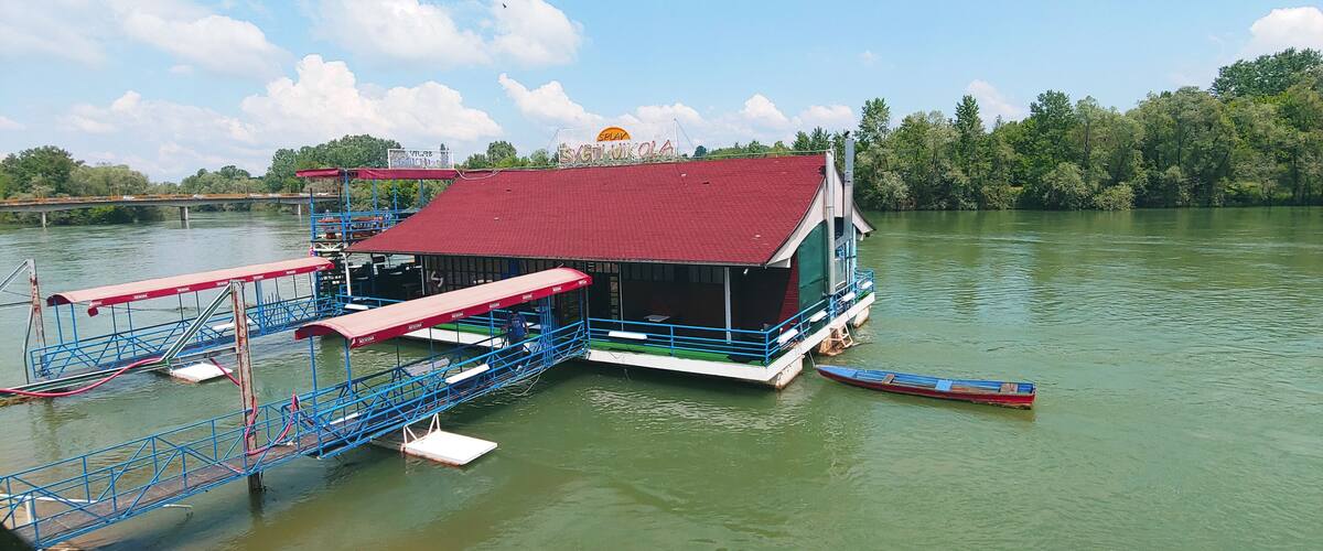 A floating house with a red roof and a sign that reads "Svetikola" on a calm river.