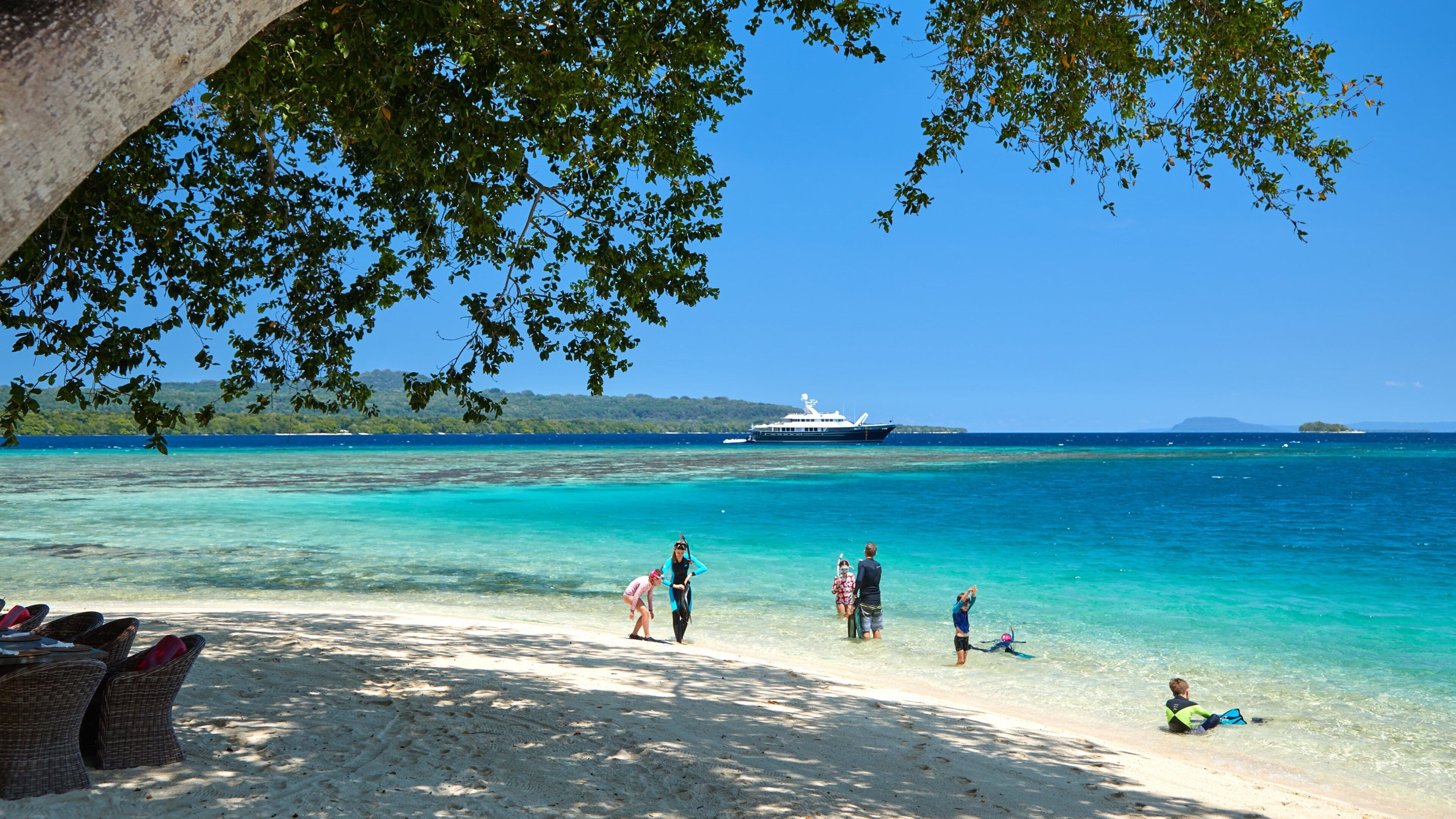 Uma praia com águas turquesa cristalinas, um iate ao longe, e pessoas aproveitando a costa.