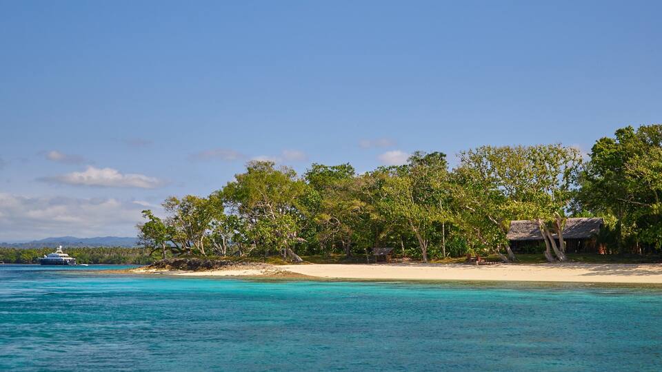 A beach with clear turquoise waters, a boat, and a thatched-roof hut surrounded by trees.