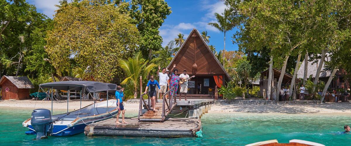 A tropical beach with a wooden pier, boats, and a thatched-roof building.