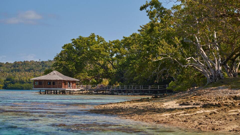A wooden house on stilts over water, surrounded by lush trees and a clear sky.
