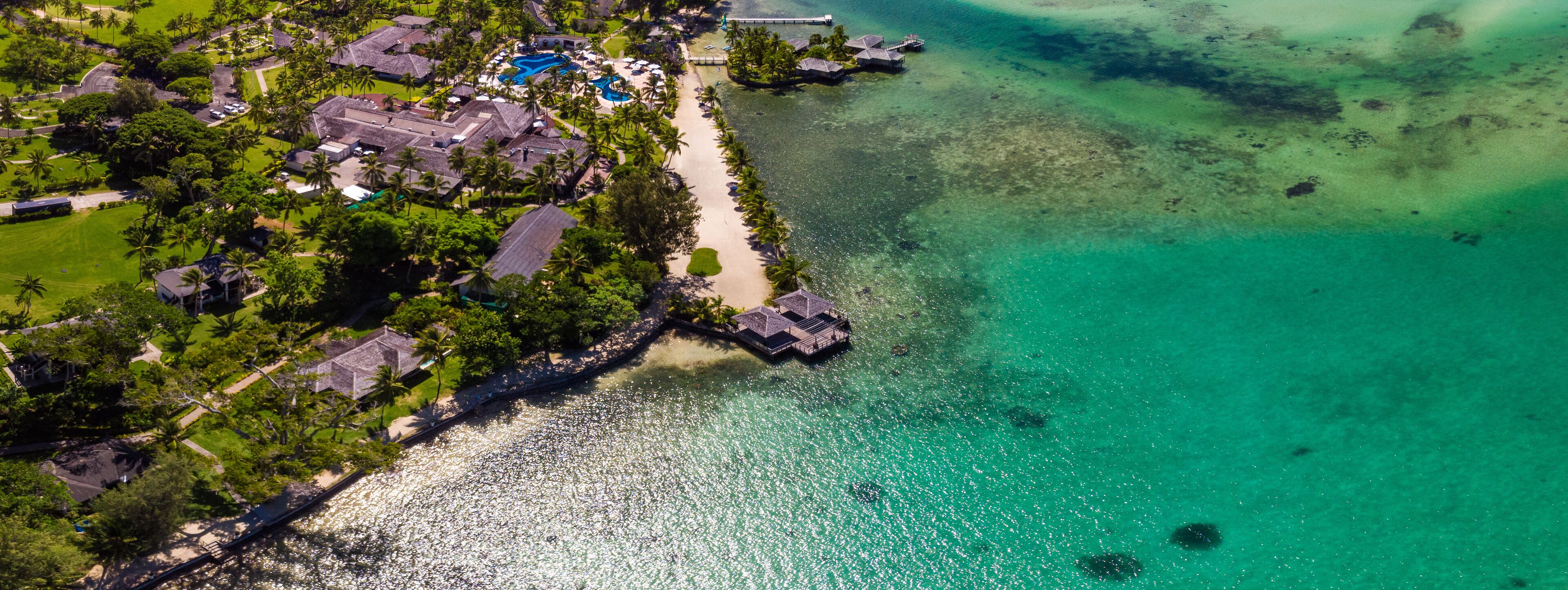Vista aérea de un complejo turístico costero con aguas turquesas cristalinas, una playa de arena y bungalows sobre el agua.