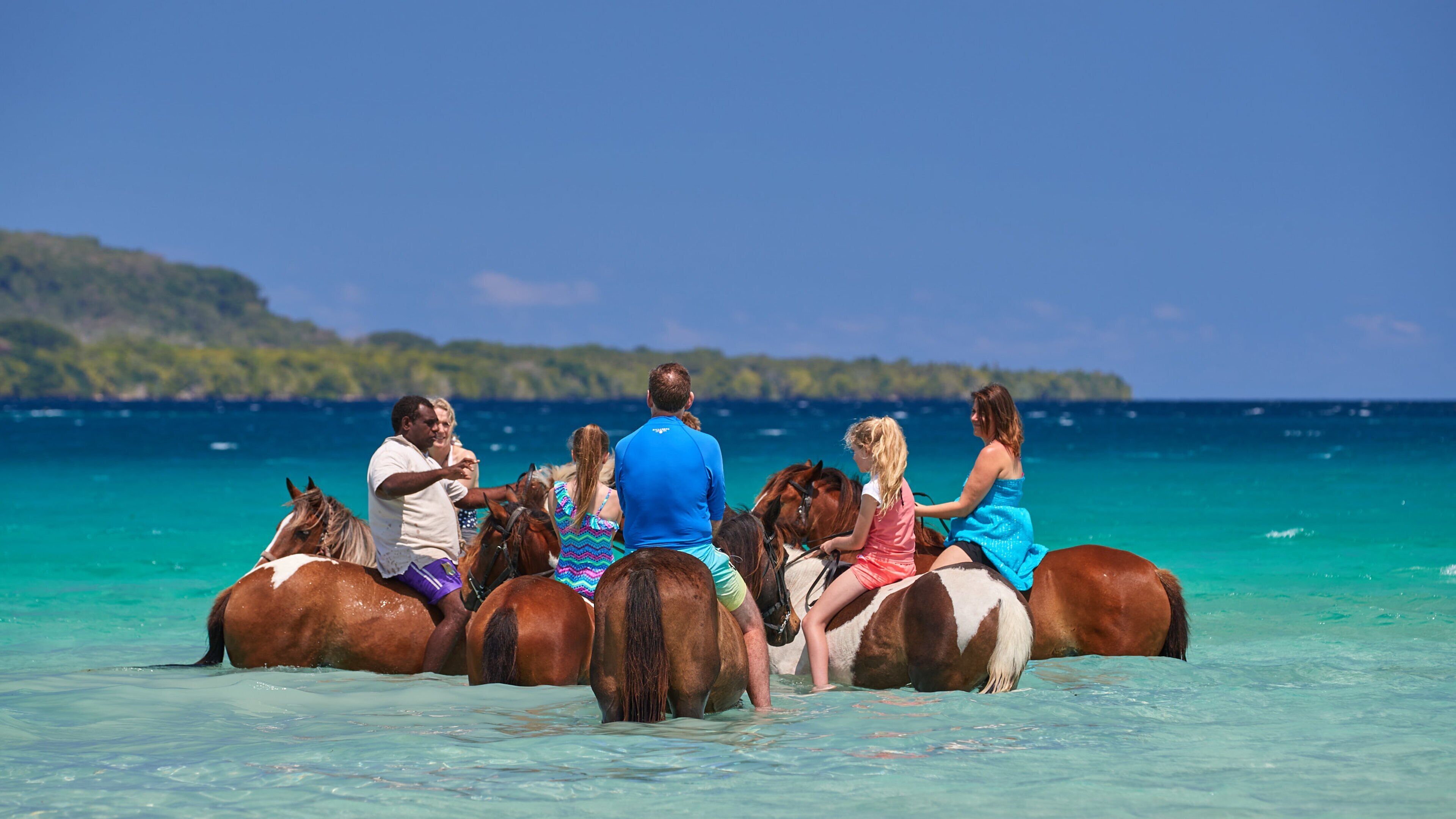 People riding horses through shallow turquoise waters with a clear blue sky and a distant island in the background.