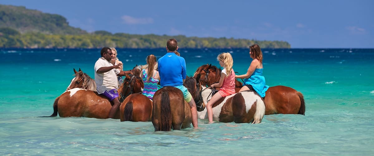 People riding horses through shallow turquoise waters with a clear blue sky and a distant island in the background.