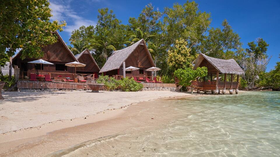 A beach with wooden huts, clear turquoise water, and lush green trees.
