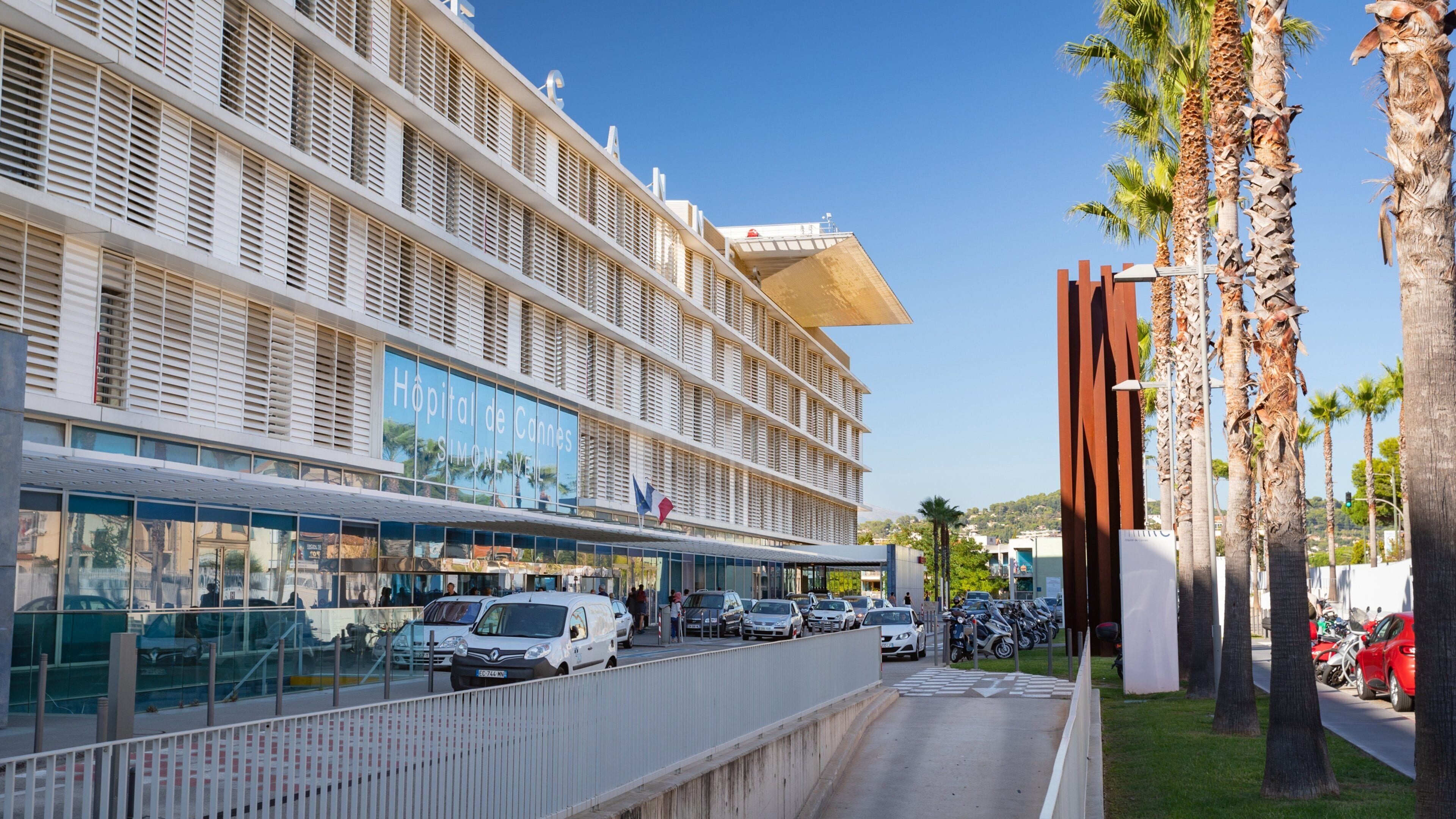 A modern hospital building with a large sign, parked cars, and palm trees.