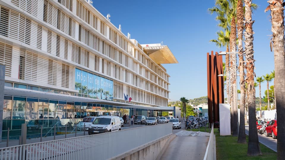Un edificio moderno de hospital con un letrero grande, coches estacionados y palmeras.