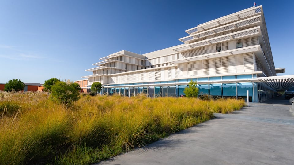Un edificio moderno con un diseño arquitectónico único, rodeado de hierba alta y un cielo azul despejado.