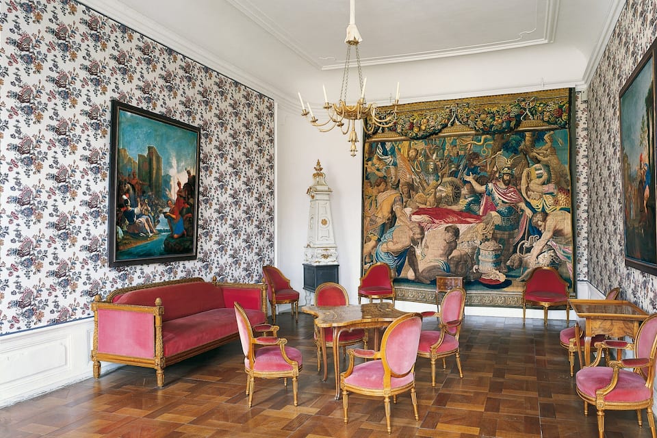 Une pièce avec un papier peint à motifs floraux, un canapé rouge, des fauteuils dorés et une grande tapisserie.