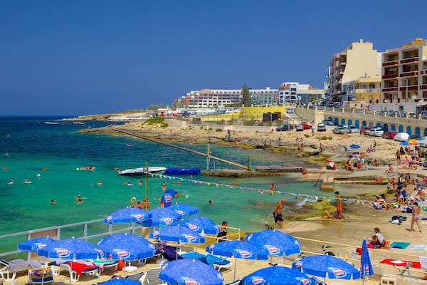 A crowded beach with clear turquoise waters, blue umbrellas, and people sunbathing.