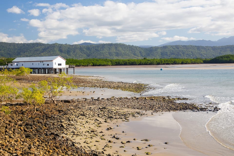 A coastal landscape with a white building on stilts, a rocky shoreline, calm water, and distant mountains.