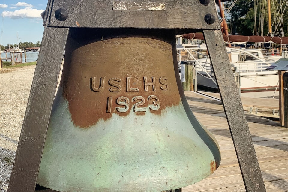 A rusted metal structure with "USLHS 1923" engraved on it, set against a backdrop of a lighthouse and marina.