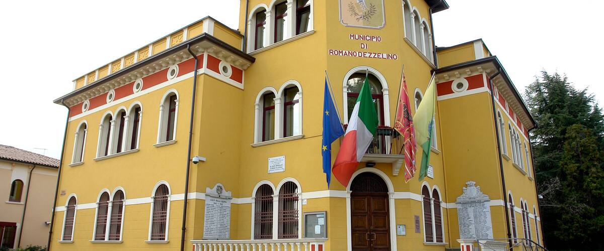 A yellow building with multiple flags, including the European Union flag, and a plaque that reads "MUNICIPIO DI ROMANO DEZZELINO".